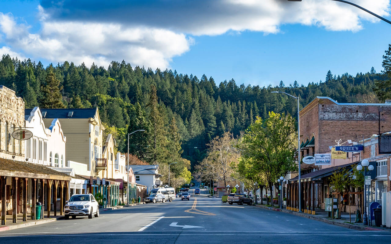 View down Calistoga's historic main street lined with shops and restaurants, backed by forested hills under a partly cloudy sky.