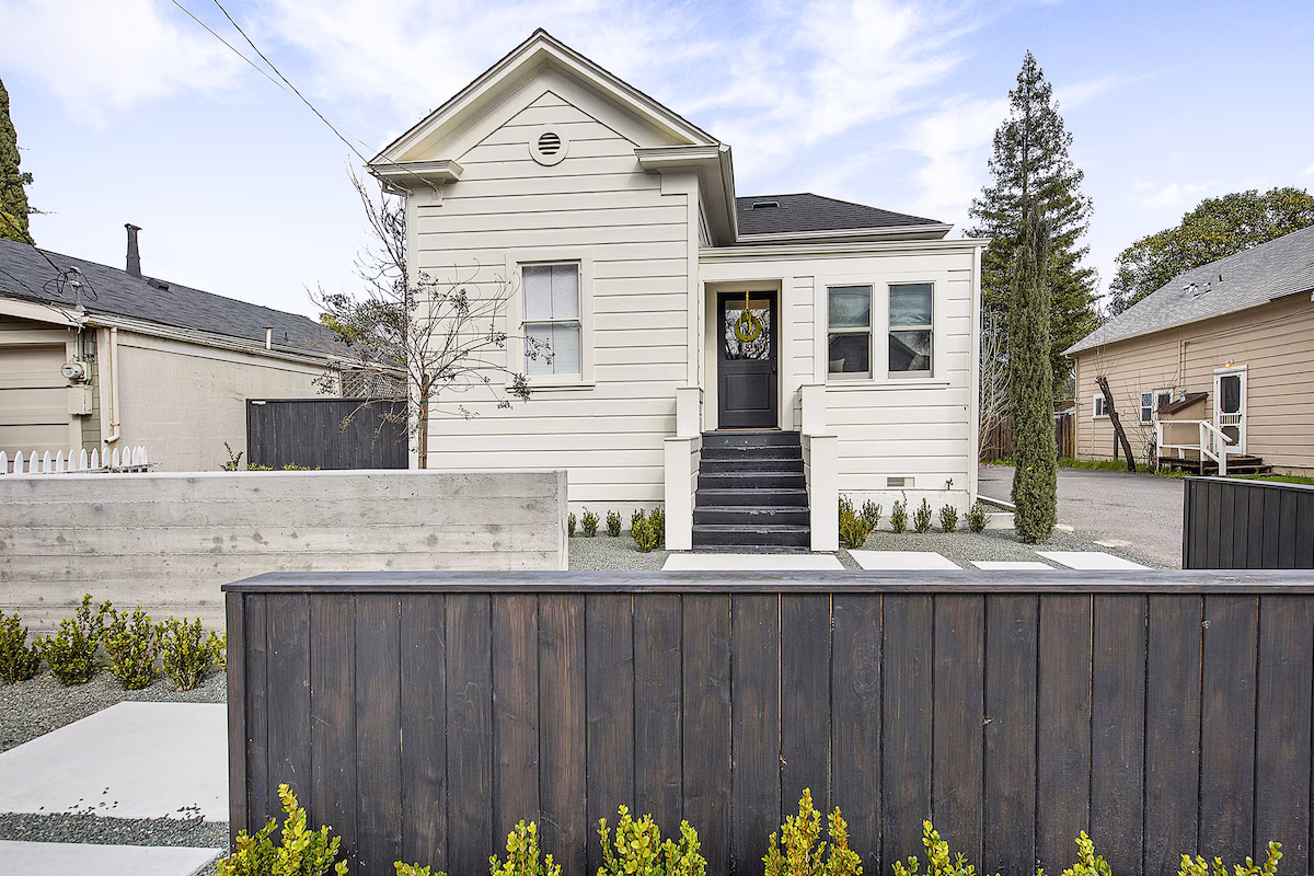 White single-story Edwardian home with gabled roof and black front door, reached by a black staircase, behind a dark wood privacy fence with landscaped greenery in front